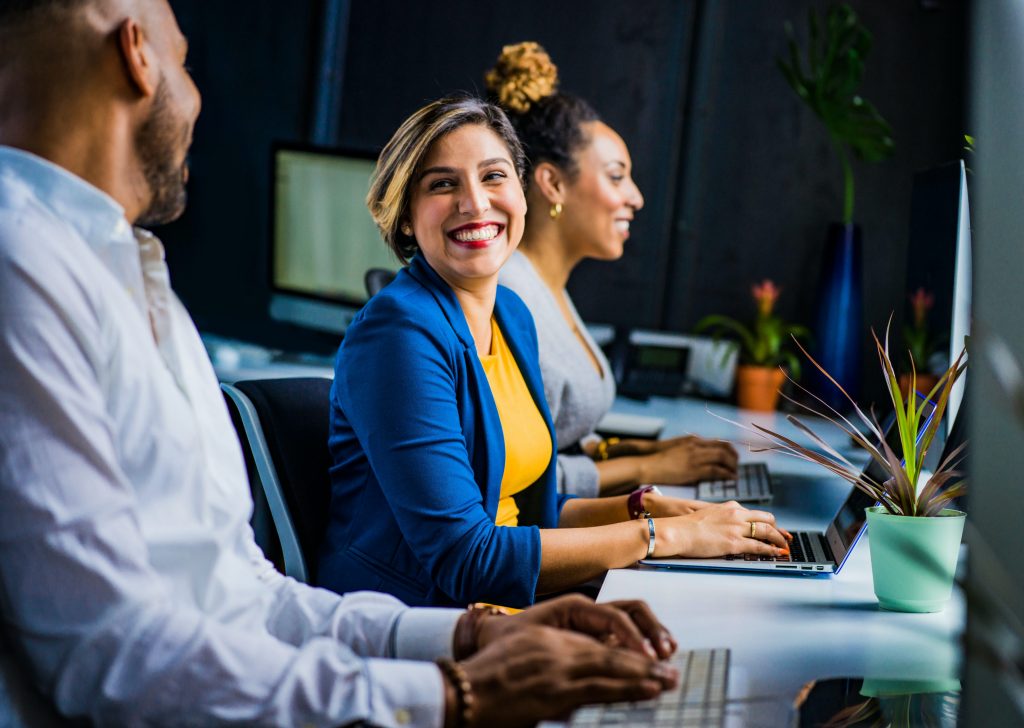 Woman smiling, sitting at a desk alongside colleagues, enjoying working in an environment with people that don't drain her.