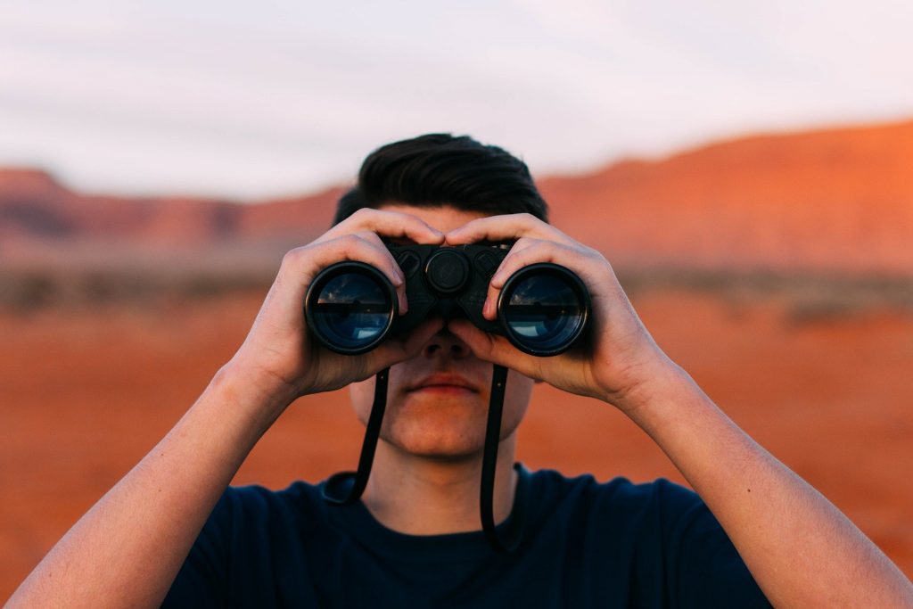 a young white man standing with a pair of binoculars standing in the middle of the desert, a vision in front of him for what he wants ahead, to help guide him