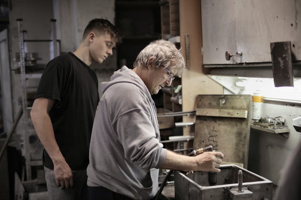 two men together in a work shop - young man serving an apprenticeship in an effort to learn more about the area he wants to move into in business