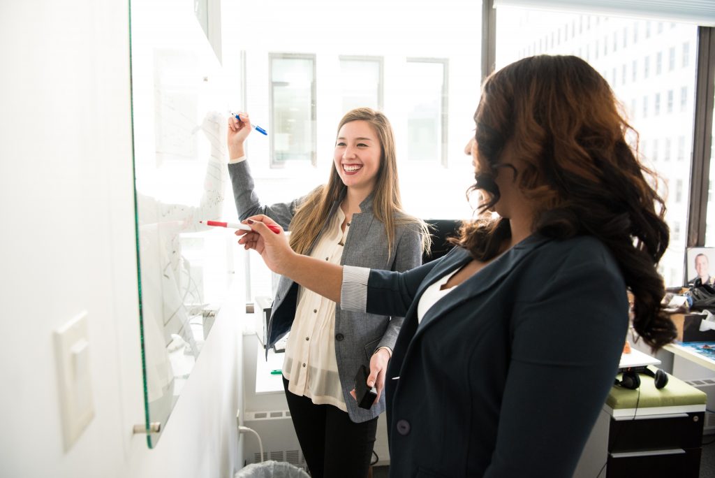 two women at whiteboard smiling, working together, wearing business attire