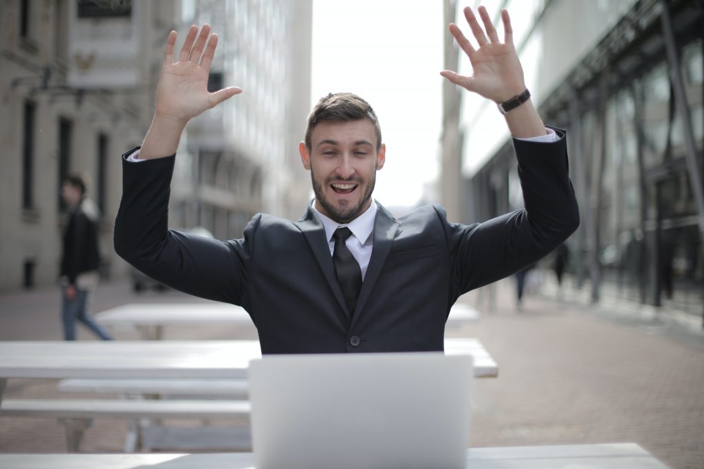businessman sitting at laptop with hands raised looking happy